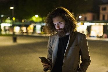 Stylish young man outdoors in the city at night looking at cell phone - MAUF00974