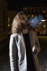 Stylish young man smoking outdoors at night - MAUF00977