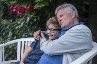 Grandfather hugging grandson on garden bench - PAF01755