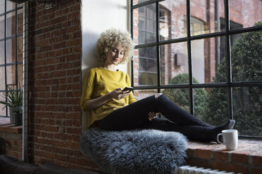 Young woman sitting on window sill, using smart phone - RBF05533