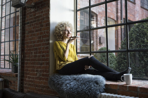 Young woman sitting on window sill, speaking into smart phone - RBF05534