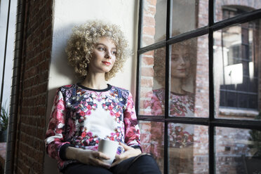 Junge Frau sitzt auf dem Fensterbrett und hält eine Tasse Kaffee in der Hand - RBF05535