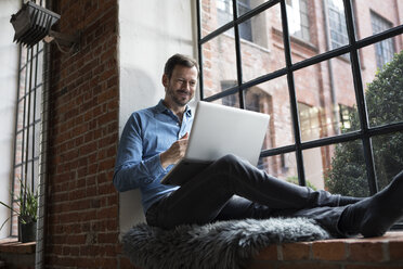 Älterer Mann sitzt auf der Fensterbank und benutzt einen Laptop - RBF05543