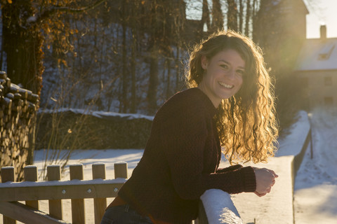 Porträt einer glücklichen jungen Frau, die sich auf ein verschneites Geländer im Gegenlicht stützt, lizenzfreies Stockfoto