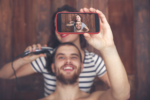 Young man taking a selfie while getting a haircut by his girlfriend with hair cutting machine - RTBF00590