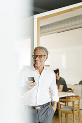 Smiling businessman with cell phone in office - PESF00416