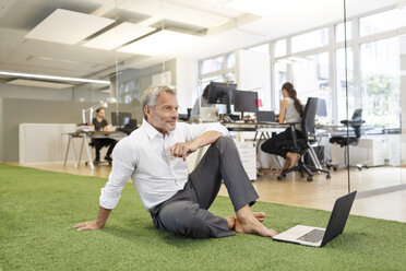 Businessman sitting on carpet in office with laptop - PESF00460