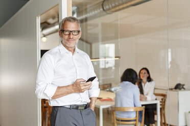 Confident businessman with cell phone in office with employees in background - PESF00475