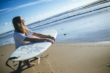 Junge Frau mit Surfbrett hockt am Strand und beobachtet den Sonnenuntergang - KIJF01083