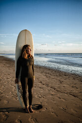 Junge Frau mit Surfbrett steht am Strand und beobachtet den Sonnenuntergang - KIJF01094