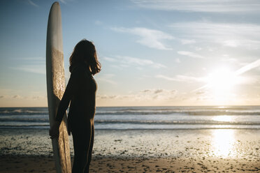 Junge Frau mit Surfbrett steht am Strand und beobachtet den Sonnenuntergang - KIJF01095