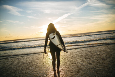 Rückenansicht einer Frau mit Surfbrett am Strand bei Sonnenuntergang - KIJF01097