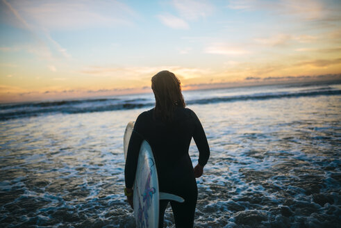 Rückenansicht einer jungen Frau mit Surfbrett, die am Meer steht und den Sonnenuntergang beobachtet - KIJF01105