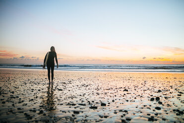 Junge Frau mit Surfbrett geht bei Sonnenuntergang am Strand spazieren - KIJF01107
