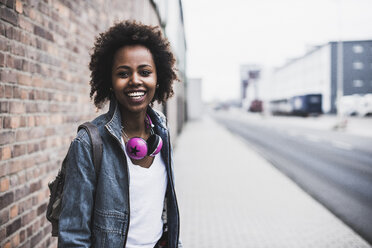 Porträt einer lächelnden jungen Frau mit Kopfhörern und Rucksack auf dem Bürgersteig stehend - UUF09784
