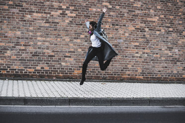 Young woman with headphones and backpack jumping in the air in front of brick wall - UUF09789