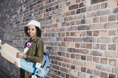 Porträt einer lächelnden jungen Frau mit Kopfhörern, Skateboard und Rucksack vor einer Backsteinmauer, lizenzfreies Stockfoto