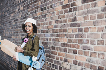 Portrait of smiling young woman with headphones, skateboard and backpack in front of brick wall - UUF09795