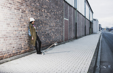 Junge Frau mit Kopfhörern, Rucksack und Skateboard lehnt an einer Backsteinmauer - UUF09796