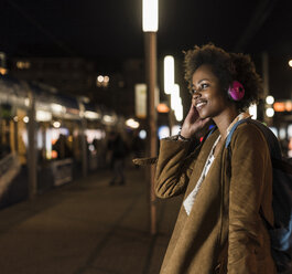 Lächelnde junge Frau, die beim Warten an der Straßenbahnhaltestelle mit Kopfhörern Musik hört - UUF09809