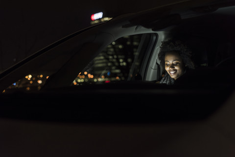 Lächelnde junge Frau, die nachts in einem Auto sitzt und ein Tablet benutzt, lizenzfreies Stockfoto