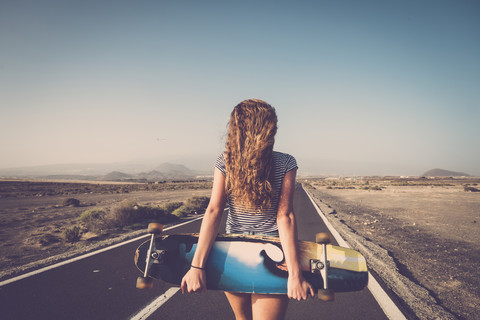 Spanien, Teneriffa, Rückenansicht einer blonden jungen Frau mit Longboard auf einer leeren Landstraße, lizenzfreies Stockfoto