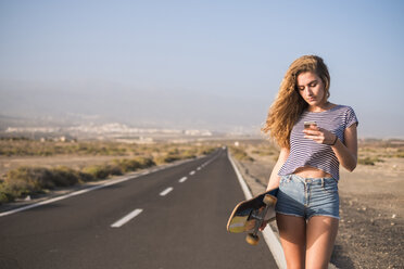 Spanien, Teneriffa, junge Frau mit Skateboard steht auf leerer Landstraße und schaut auf ihr Smartphone - SIPF01341