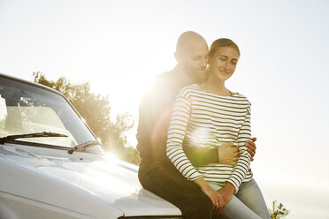 Young couple in love sitting on car bonnet at backlight - SRYF00224