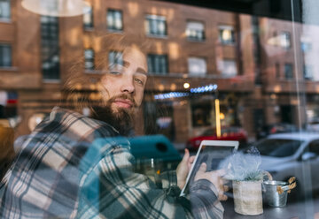 Porträt eines stilvollen jungen Mannes hinter einer Fensterscheibe mit Tablet in einem Café - MGOF02814