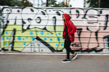 Young man wearing red hoodie passing a graffiti wall - JRF01161