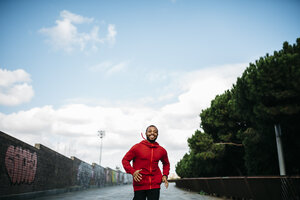 Smiling young man wearing red hoodie running in the city - JRFF01168