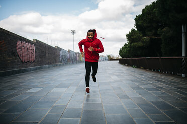 Smiling young man wearing red hoodie running in the city - JRF01169