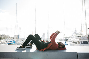 Young man lying on a wall at the marina using cell phone - JRFF01171