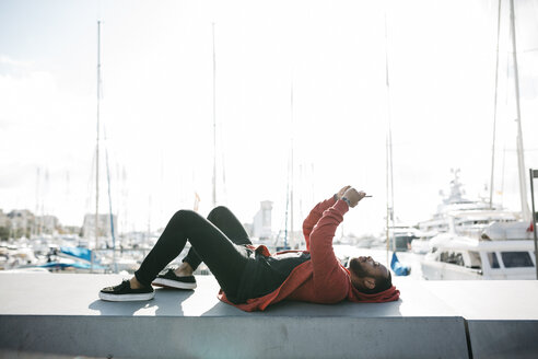 Junger Mann liegt auf einer Mauer im Jachthafen und benutzt ein Mobiltelefon - JRFF01171