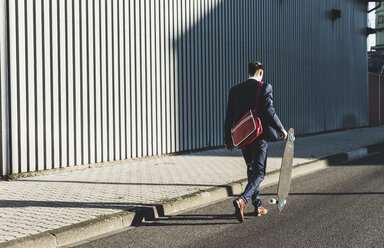 Junger Geschäftsmann zu Fuß mit Skateboard auf der Straße - UUF09823