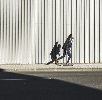 Junger Mann mit Gitarrenkoffer fährt Skateboard auf dem Bürgersteig - UUF09832