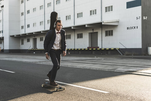 Junger Mann mit Gitarrenkoffer fährt Skateboard auf der Straße - UUF09835