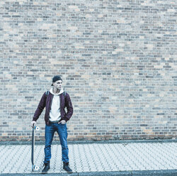 Serious young man with skateboard in front of brick wall - UUF09855