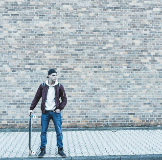 Ernster junger Mann mit Skateboard vor einer Backsteinmauer - UUF09855