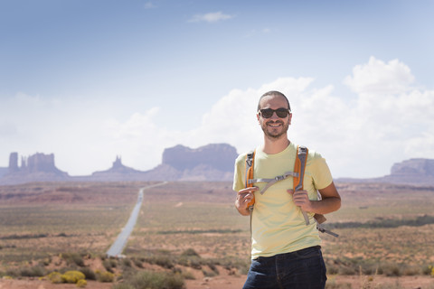 USA, Utah, Porträt eines lächelnden Mannes mit Rucksack und Sonnenbrille auf dem Weg zum Monument Valley, lizenzfreies Stockfoto