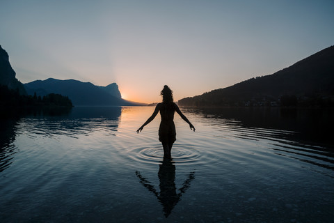 Österreich, Mondsee, Mondsee, Silhouette einer im Wasser stehenden Frau bei Sonnenuntergang, lizenzfreies Stockfoto