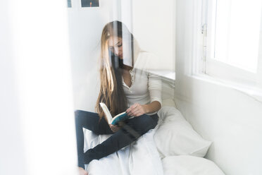 Young woman sitting on bed reading book - KKAF00316