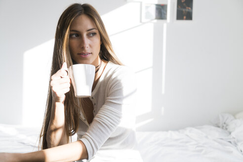 Junge Frau sitzt auf dem Bett und hält eine Tasse Kaffee - KKAF00317