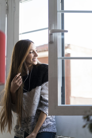 Lächelnde junge Frau mit Kopfhörern, die aus dem Fenster schaut, lizenzfreies Stockfoto