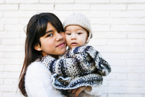 Porträt eines lächelnden Mädchens, das ein Baby an einer Backsteinmauer hält - VABF01035
