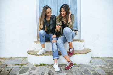 Zwei junge Frauen sitzen auf der Treppe und schauen auf ihr Handy - KIJF01132