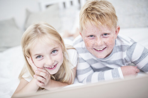 Porträt eines lächelnden Geschwisterpaares, das mit einem Laptop im Bett liegt - WESTF22539
