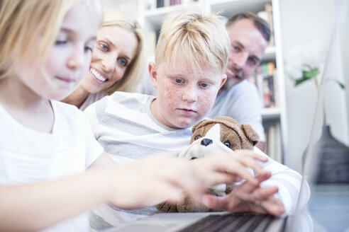 Bruder und Schwester mit Kuscheltier benutzen gemeinsam einen Laptop und werden dabei von ihren Eltern beobachtet - WESTF22556