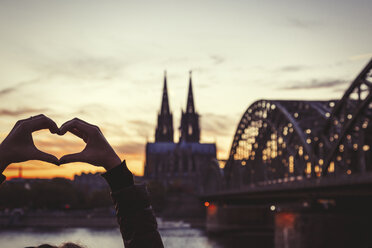 Deutschland, Köln, Frau formt Herz mit ihren Händen vor dem Kölner Dom und der Hohenzollernbrücke in der Abenddämmerung - JUNF00775