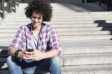 Porträt eines lächelnden jungen Mannes, der auf einer Treppe sitzt und auf sein Mobiltelefon schaut - ABZF01794